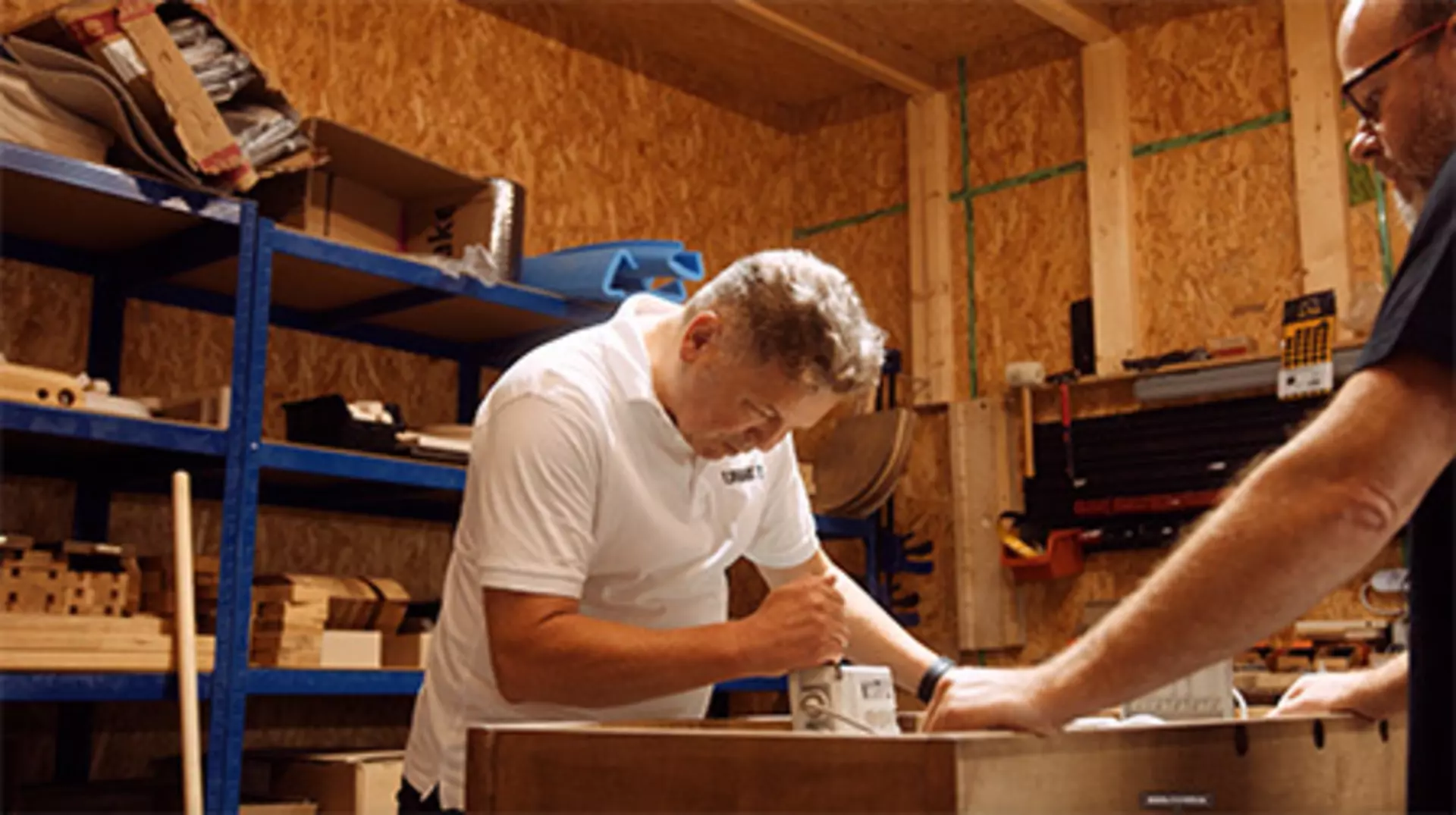 Two men in a workshop with blue shelves. Man in white t-shirt looks at a measuring device.