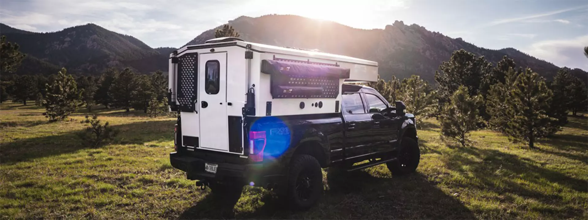 A truck camper, with a roof system using LINAK actuators, parked on grass in a scenic landscape.