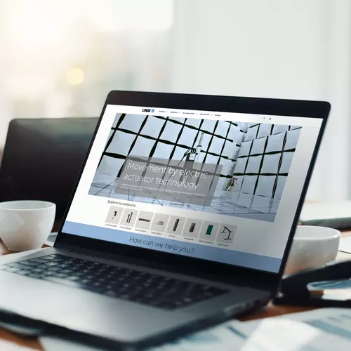 Laptop showing the LINAK website on a desk with coffee, phone, and documents.