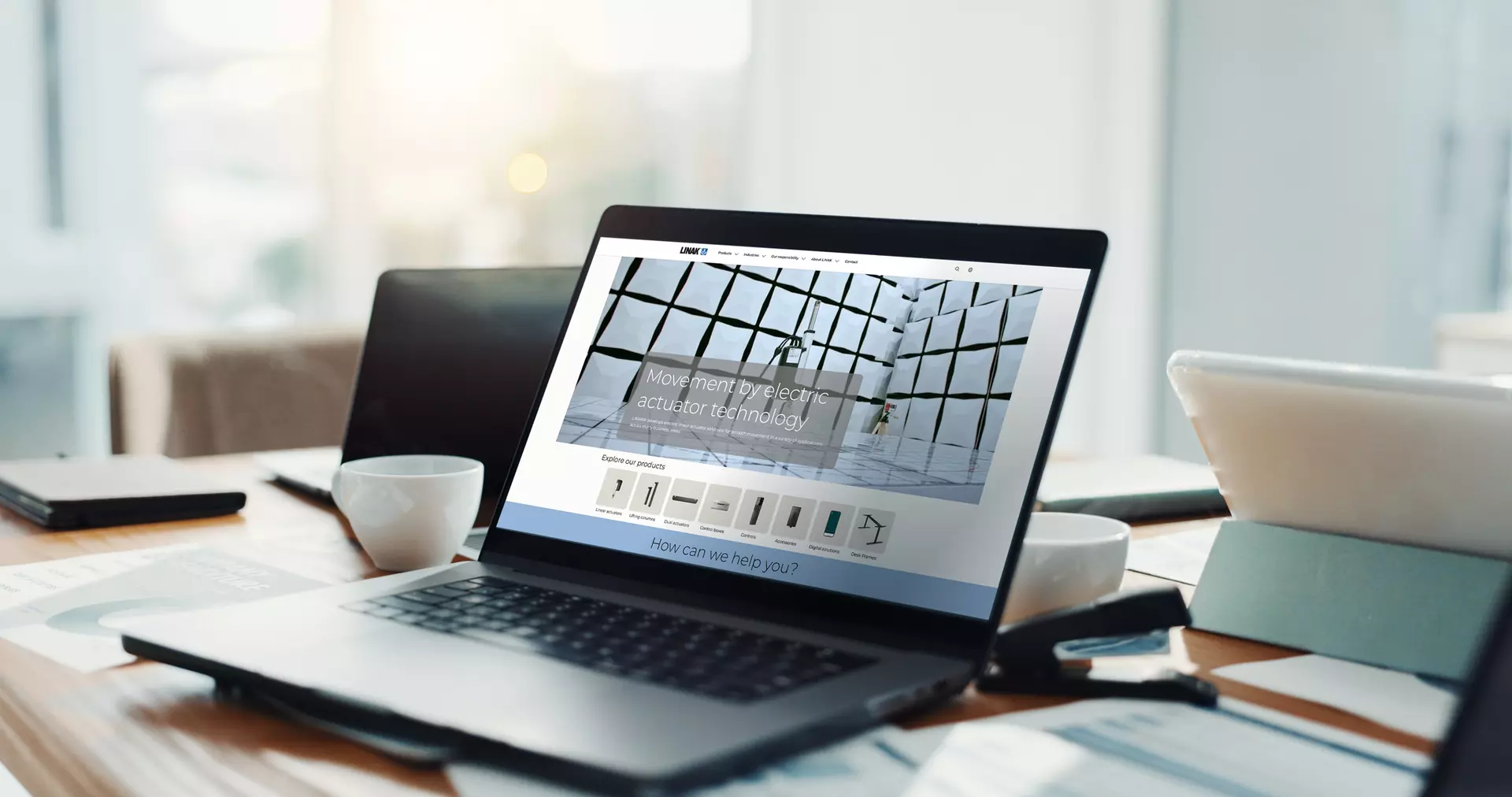 Laptop showing the LINAK website on a desk with coffee, phone, and documents.